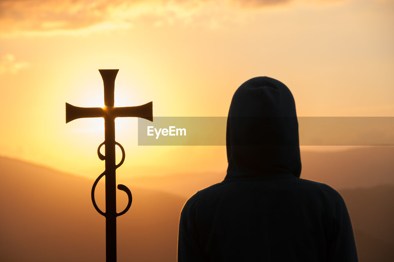 Dark silhouette of person in a hood and christian cross against sun on the background