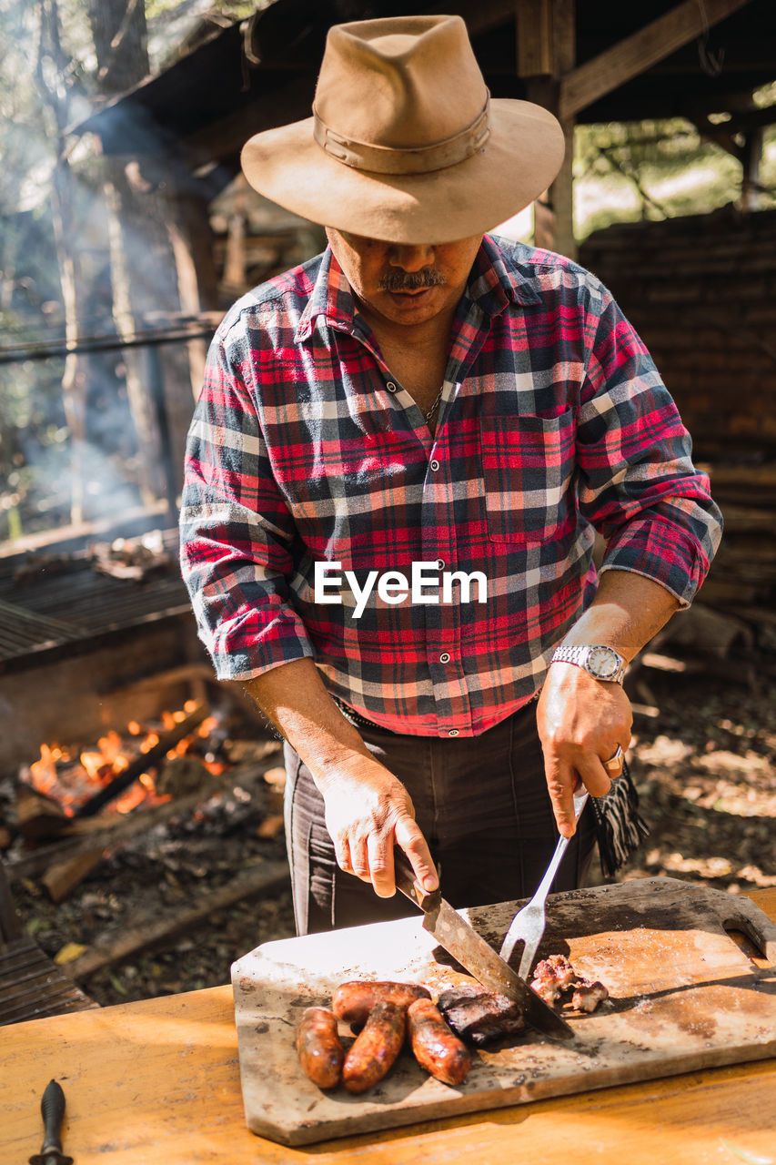 Argentinian male in hat with knife and carving fork cutting yummy meat piece on board against barbecue rack in sunlight