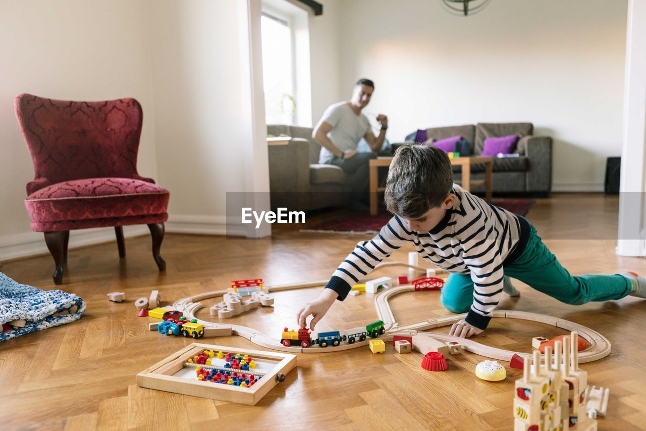 Son playing with miniature train while father sitting on sofa in background at home