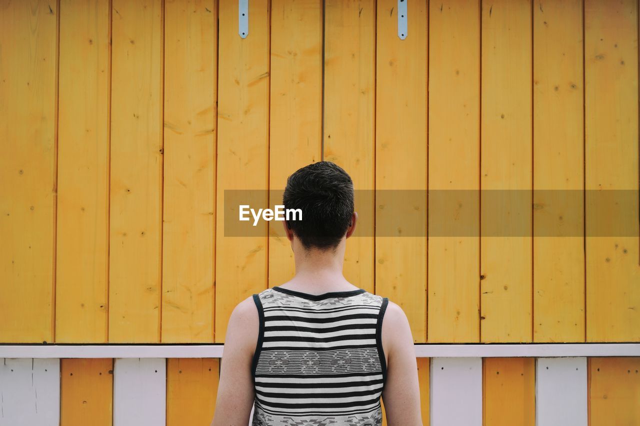 Rear view of man standing against yellow wooden wall