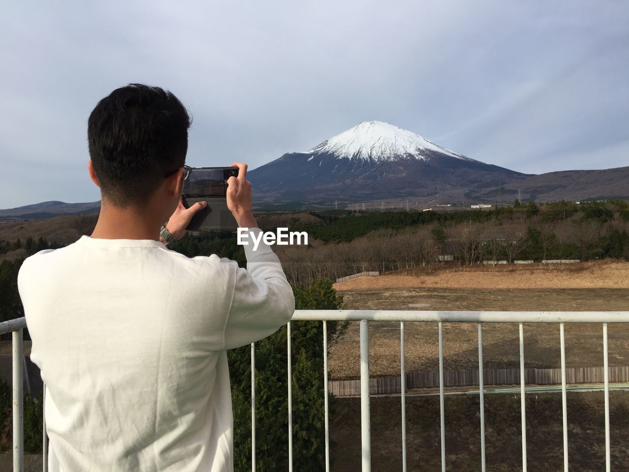 Rear view of man photographing with mobile phone while standing by railing against mountains and sky