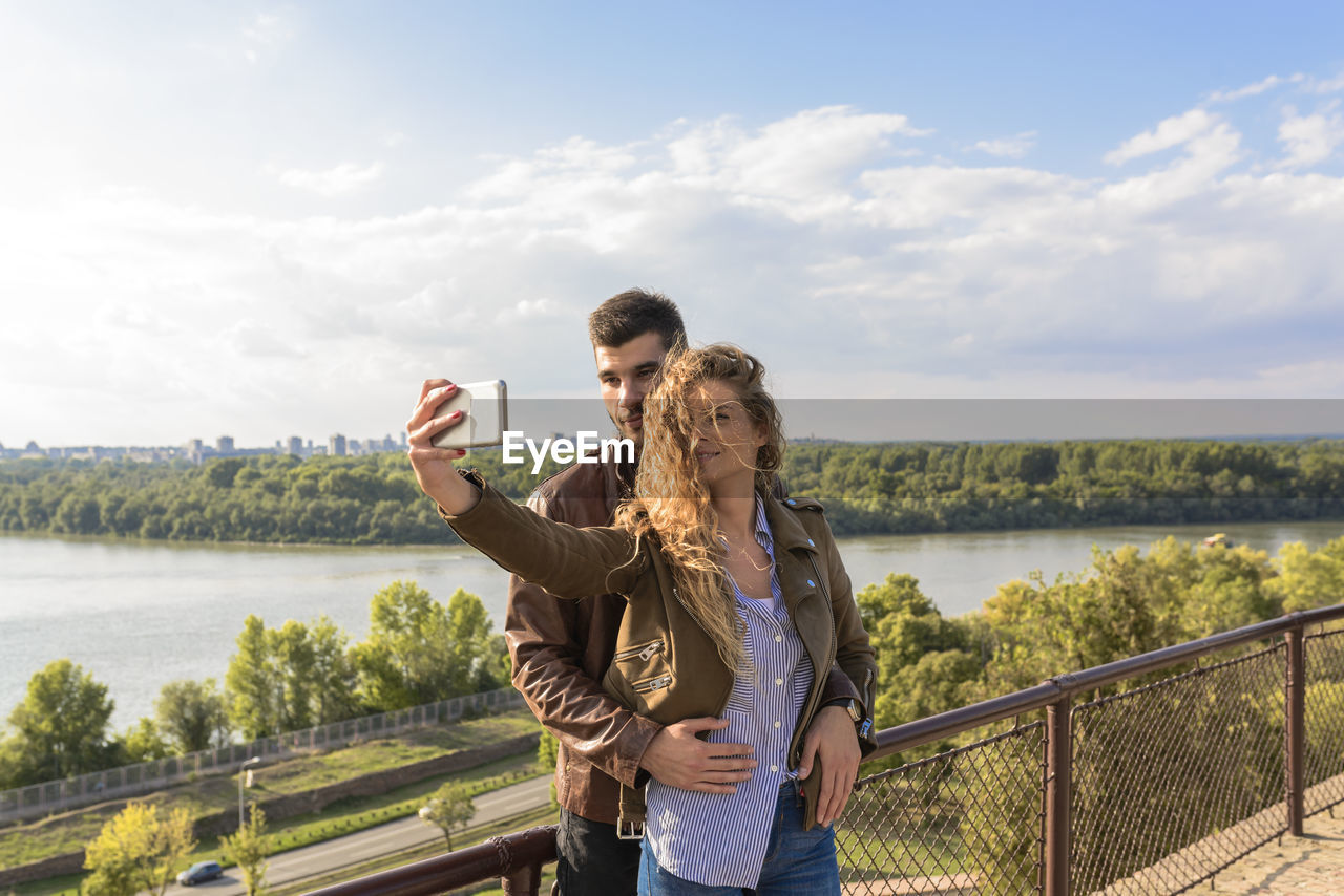 Smiling young couple taking selfie with mobile phone while standing against river