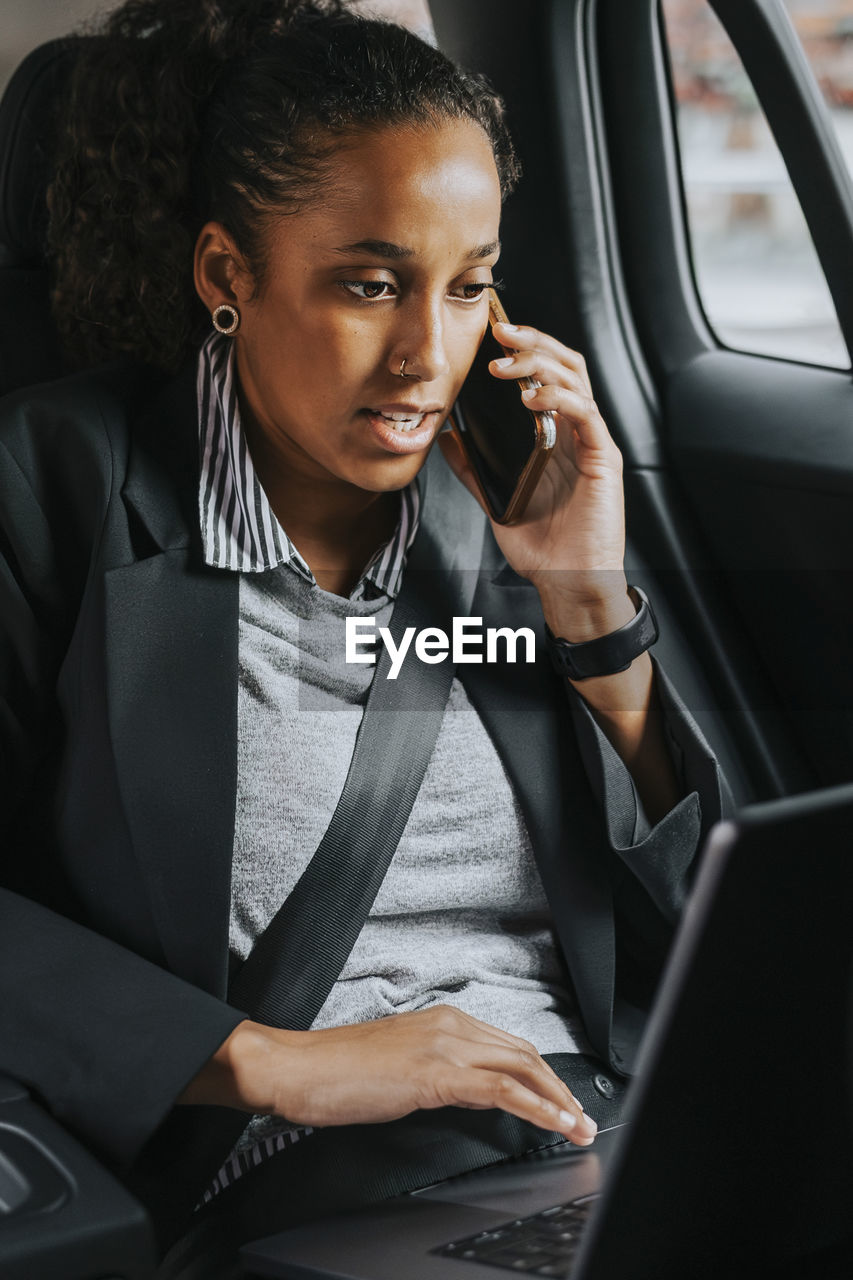 Businesswoman talking on smart phone while using laptop sitting in car