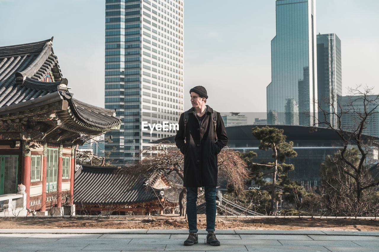 Young man in a hat, black coat and jeans stands on the street. skyscrapers and asian temple