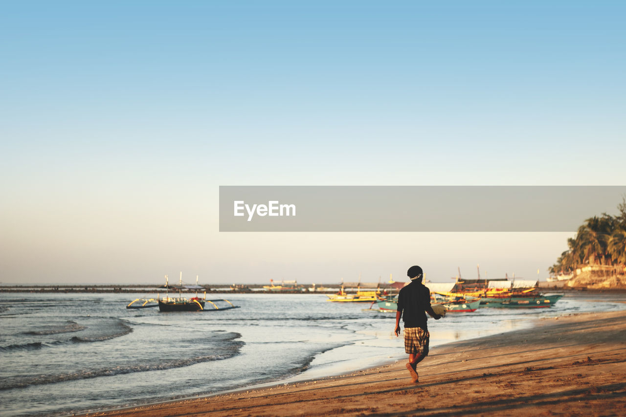 Rear view of man walking on beach by anchored boats