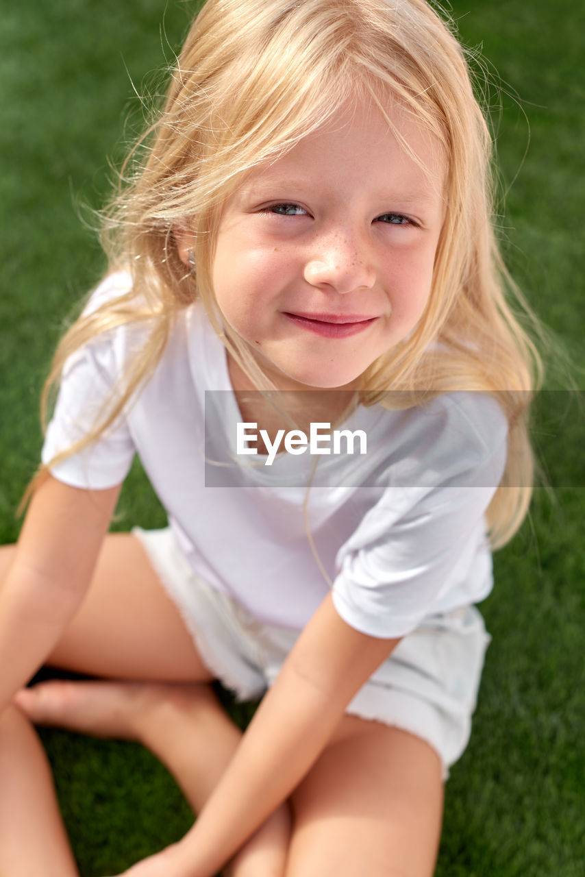 Portrait pretty smiling blonde girl sitting on green grasson field mock up white t shirt top view.