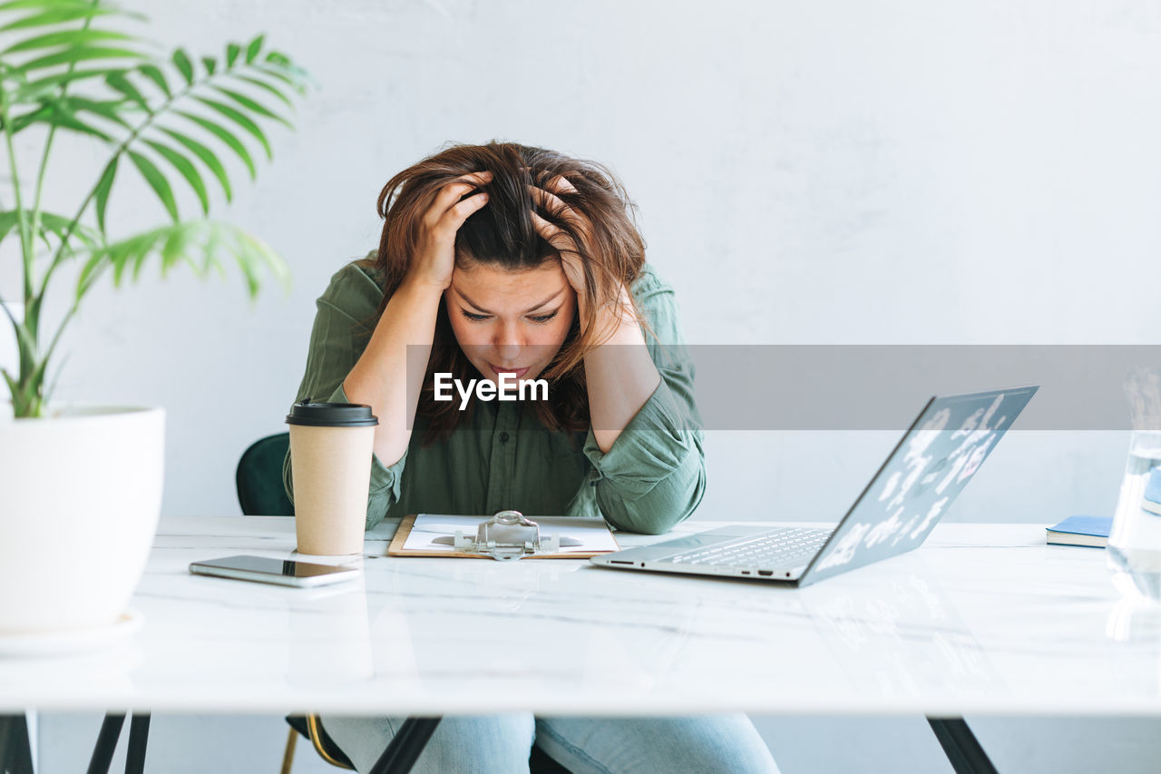 Young unhappy brunette woman plus size working at laptop on table with house plant in modern office