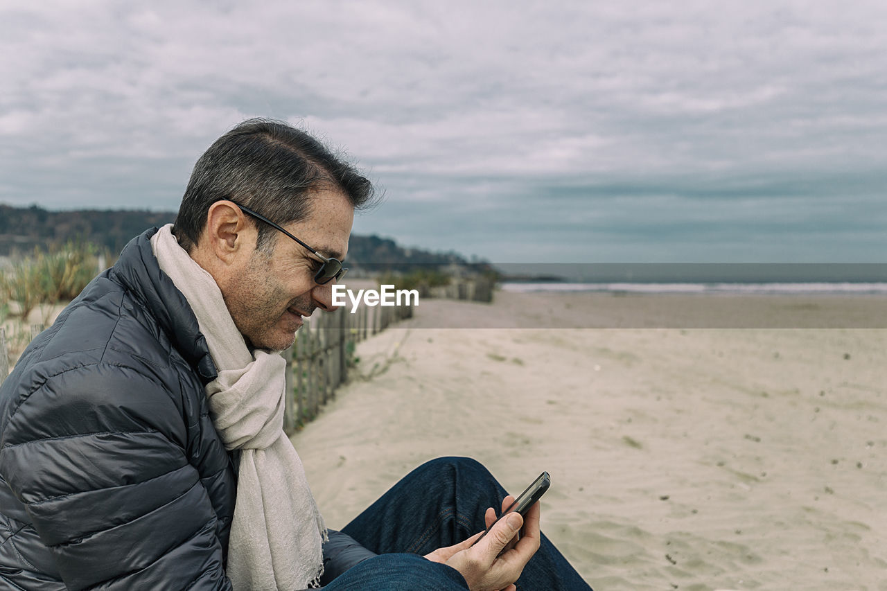 Man using phone while sitting at beach against sky