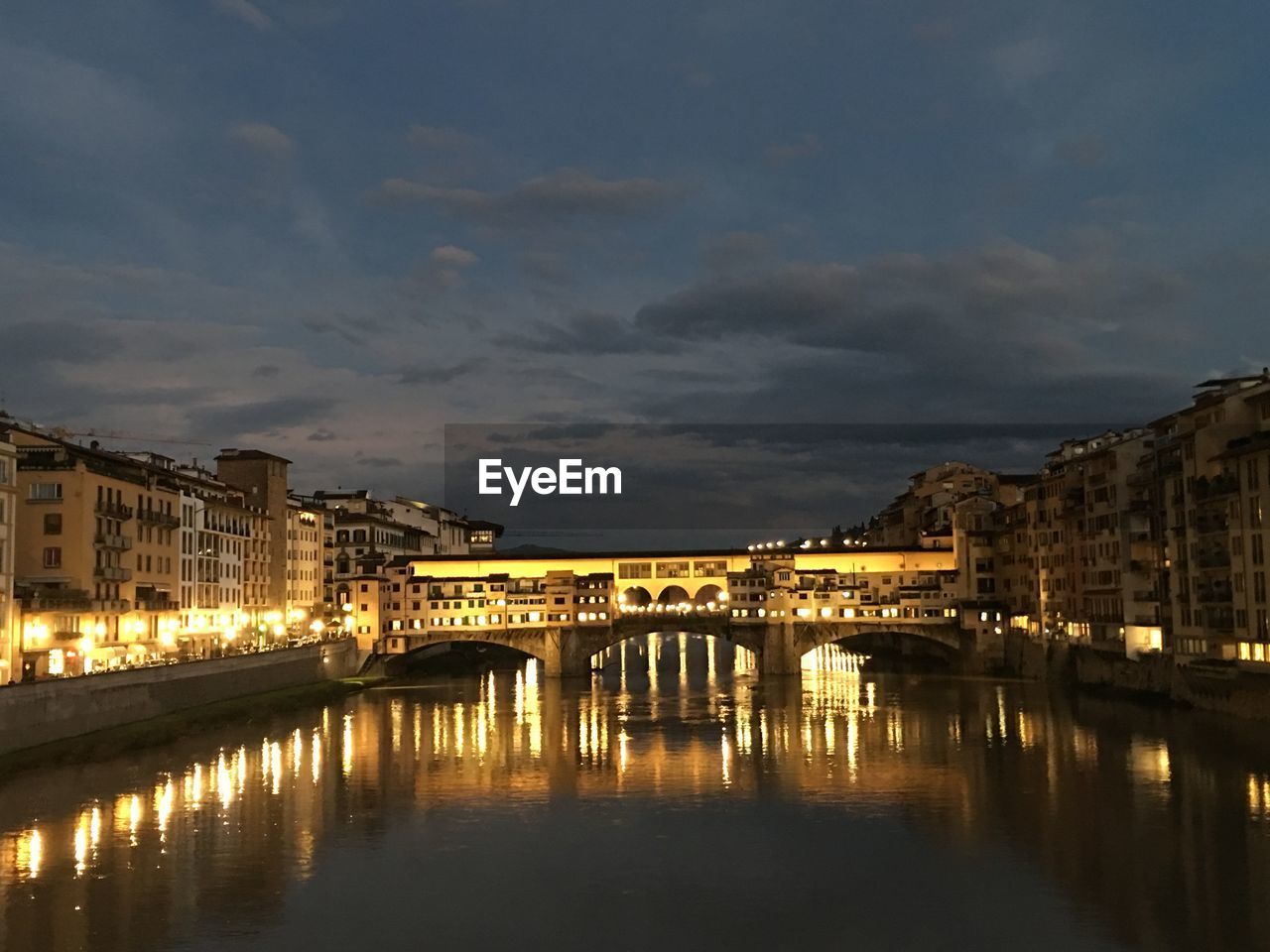 Illuminated bridge over river against cloudy sky