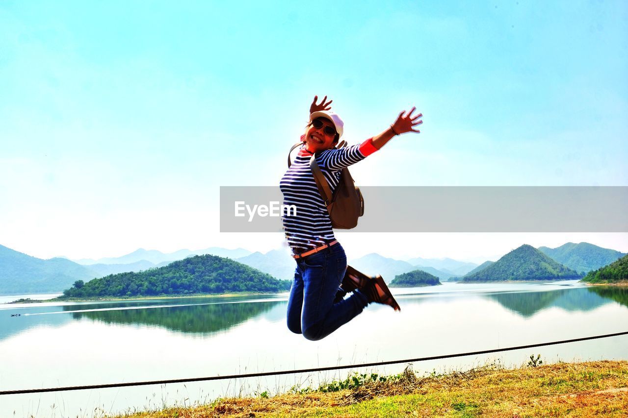Portrait of happy young woman jumping by lake against clear sky during sunny day