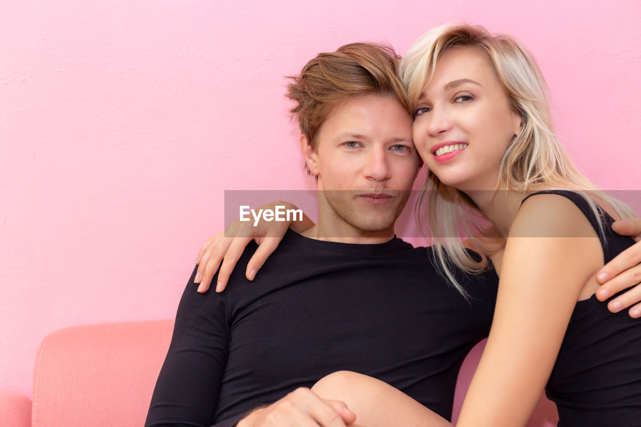 Portrait of couple sitting on sofa against pink wall