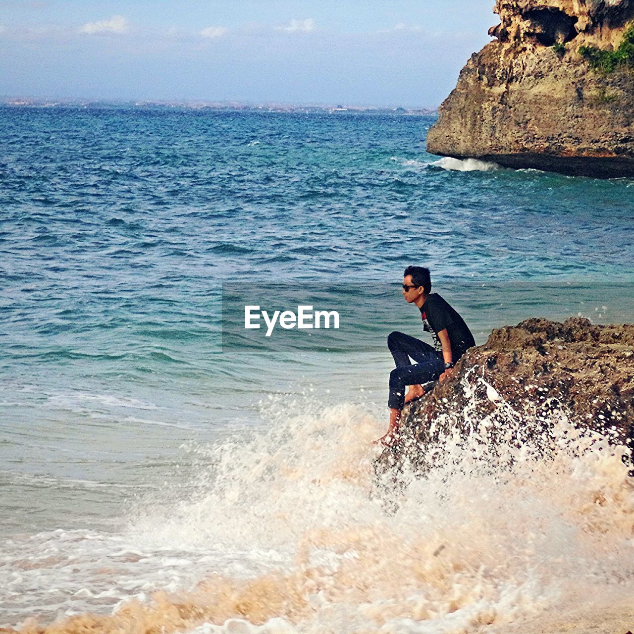 Side view of young man sitting on rock at beach against sky