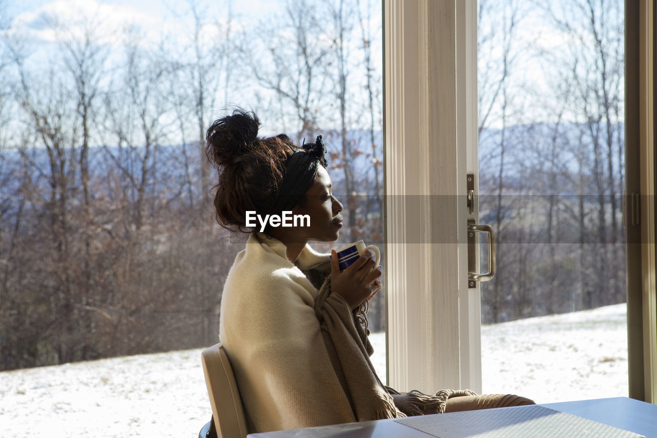 Side view of woman wrapped in shawl with eyes closed sitting by window at home