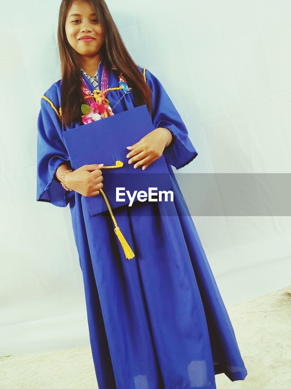 Portrait of young woman in graduation gown standing against wall