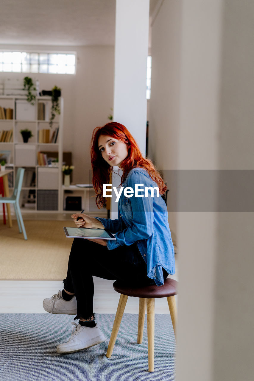 Redhead businesswoman using digital tablet while sitting on stool at office