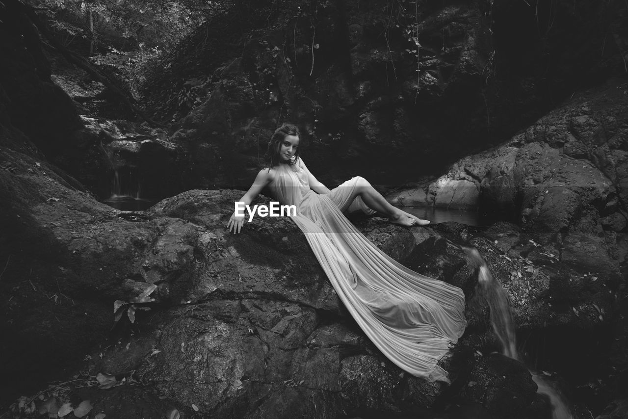 Portrait of young model wearing dress while sitting at waterfall