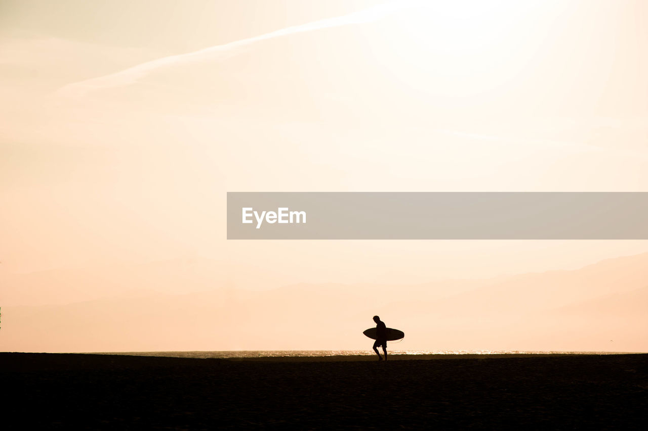 Silhouette man carrying skateboard walking on land against sky during sunset