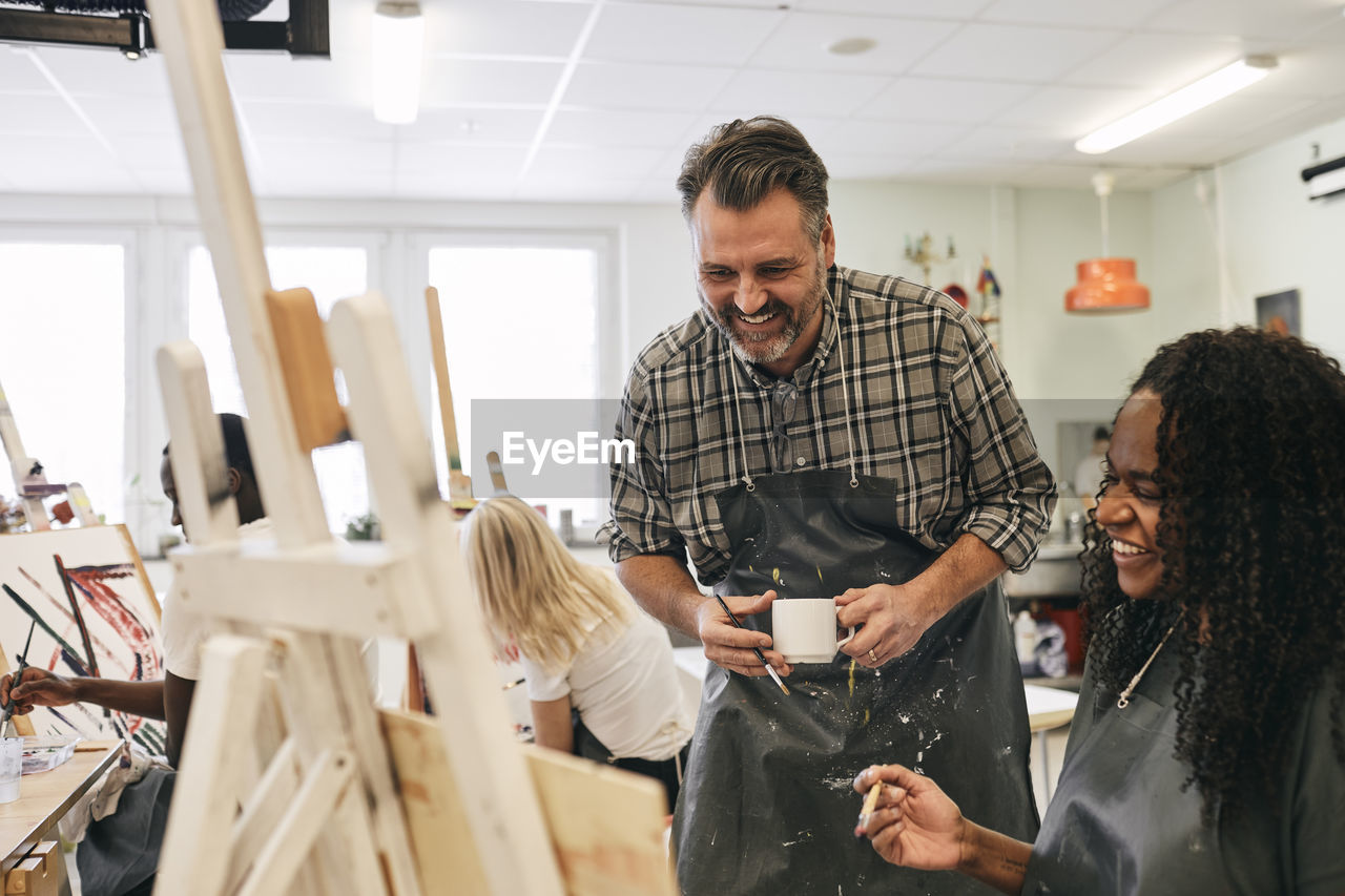 Smiling mature male tutor looking at artist's canvas by student sitting in art class