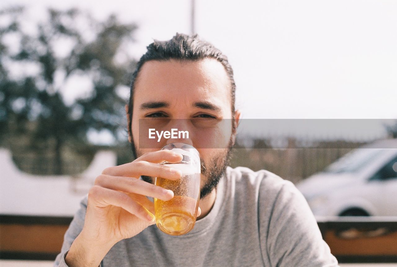 Portrait of young man drinking beer while sitting against sky