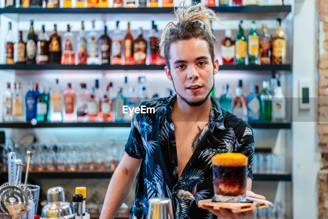 Stylish male bartender with dreadlocks standing at counter in bar with cold alcohol cocktail and looking at camera