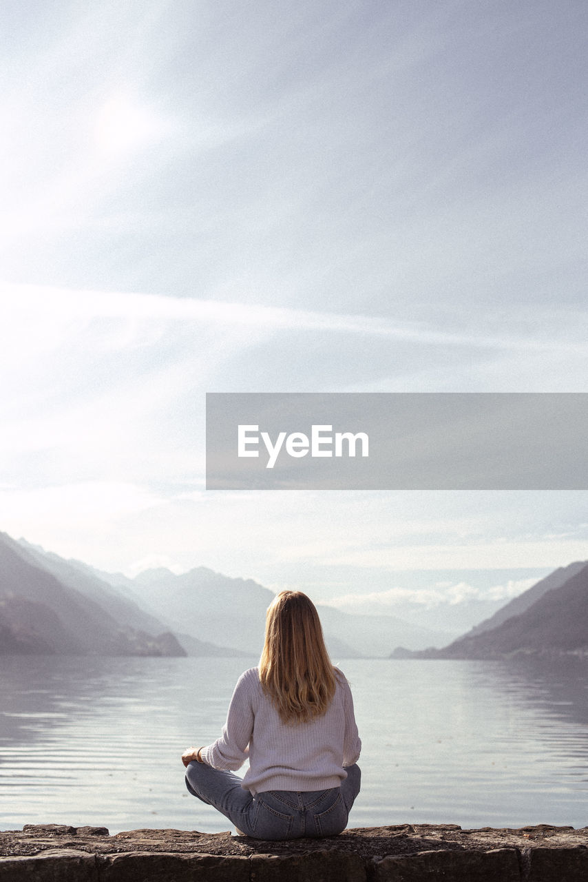 Rear view of woman sitting by lake against sky