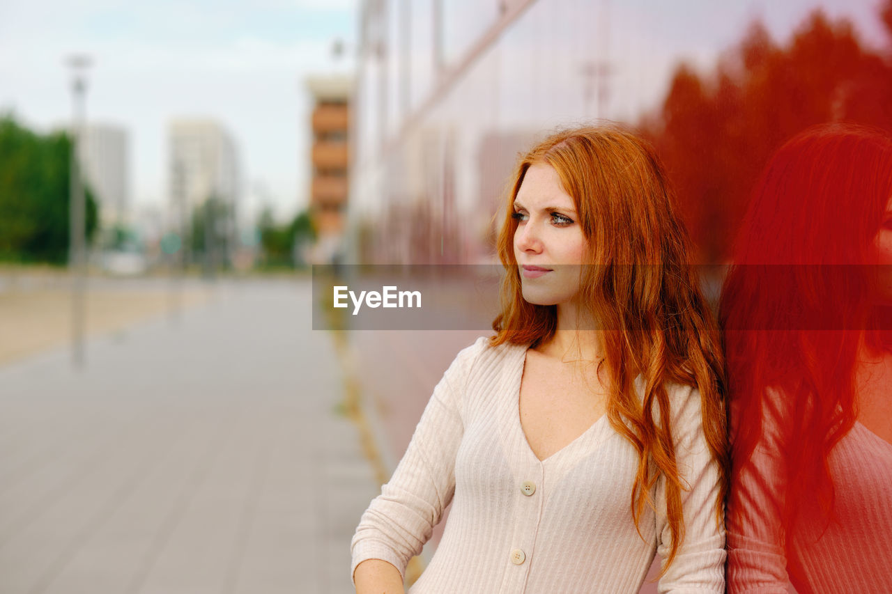 Young redhead female in elegant beige dress standing on pavement against bright red wall of modern urban building