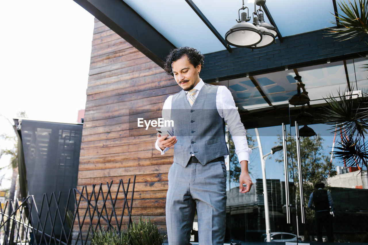 Serious bearded male entrepreneur in formal suit browsing cellphone while standing on veranda of modern building on street in city