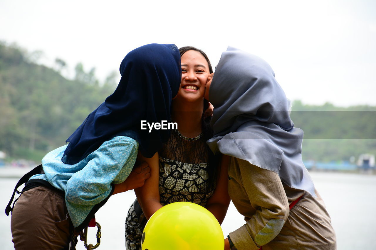 Women kissing smiling friend standing against lake