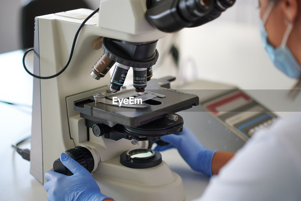 Crop unrecognizable female specialist in uniform with medical sample near microscope at work in laboratory