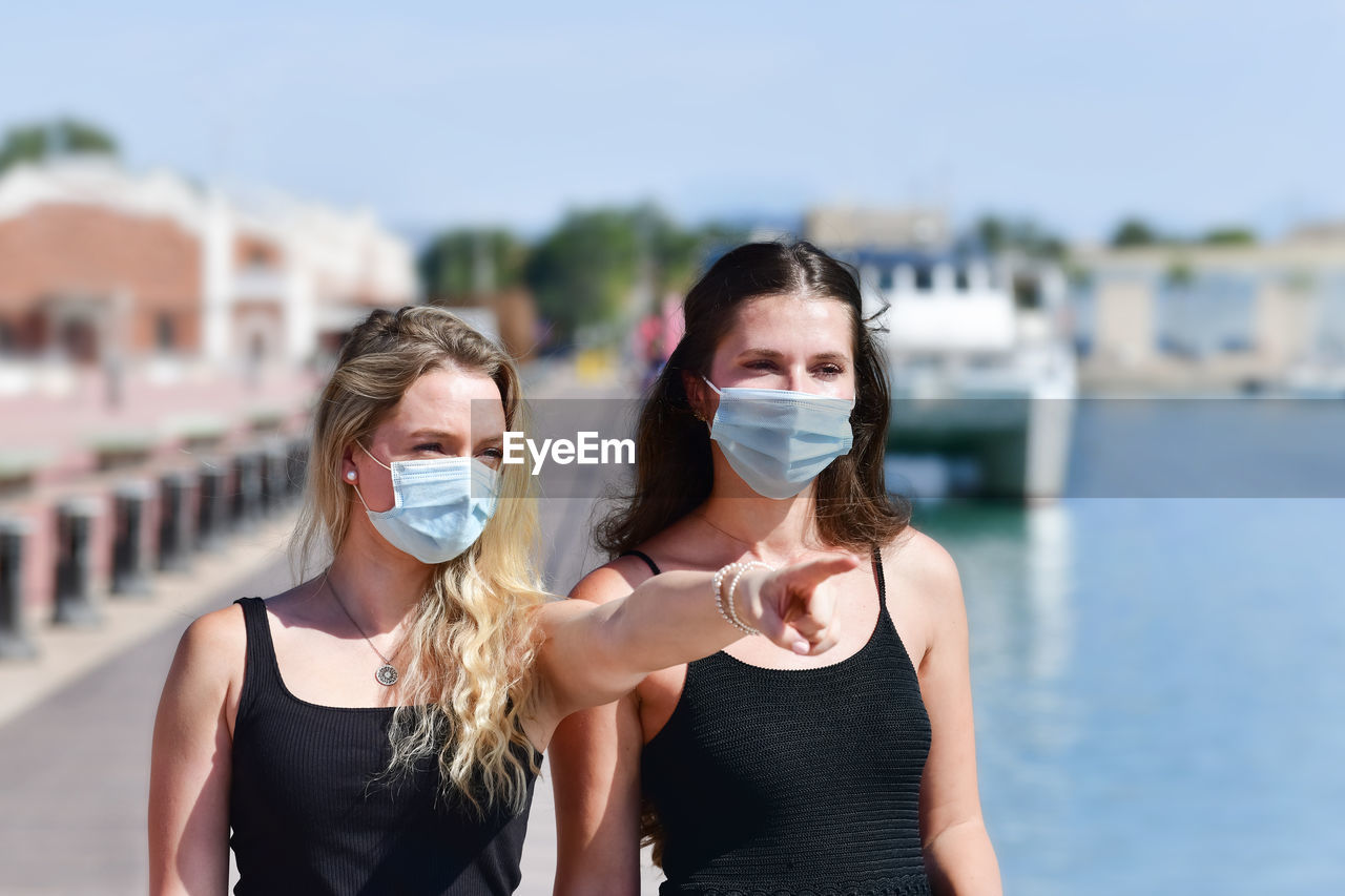 Portrait of young woman wearing mask pointing away while standing with friend at harbor