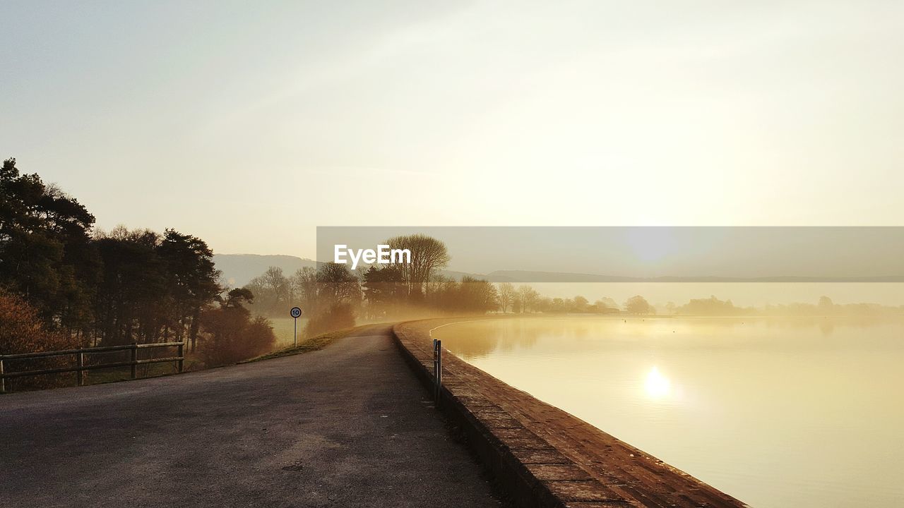 Empty road by calm lake against sky during sunrise