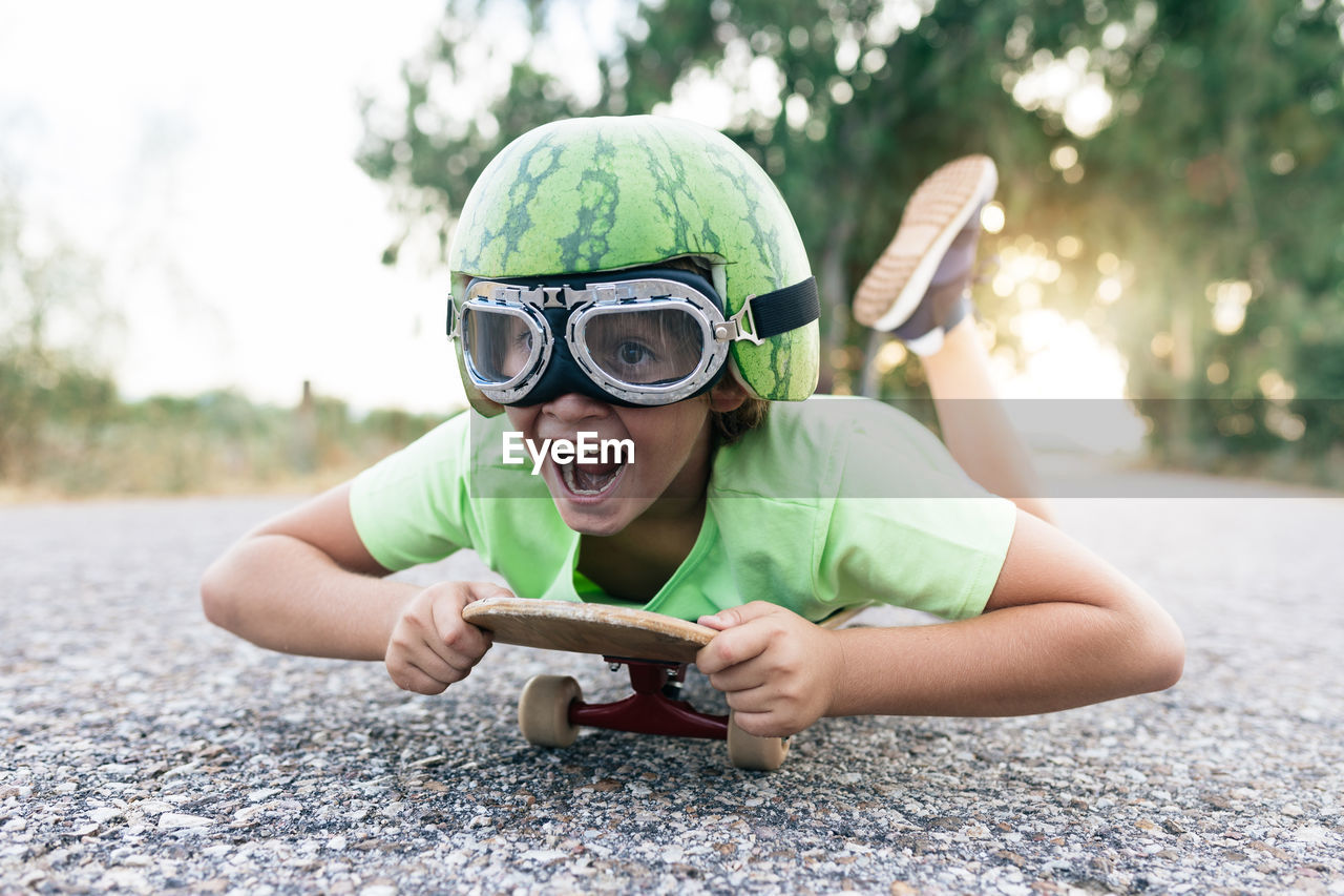 Ground level of happy kid in protective eyewear and ornamental watermelon helmet lying on skateboard on roadway and looking away