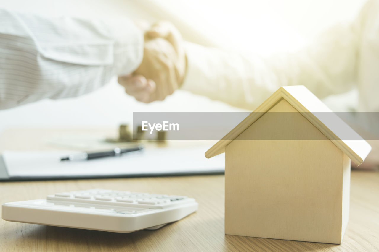 Close-up of model home and calculator on table against businessmen shaking hands in background