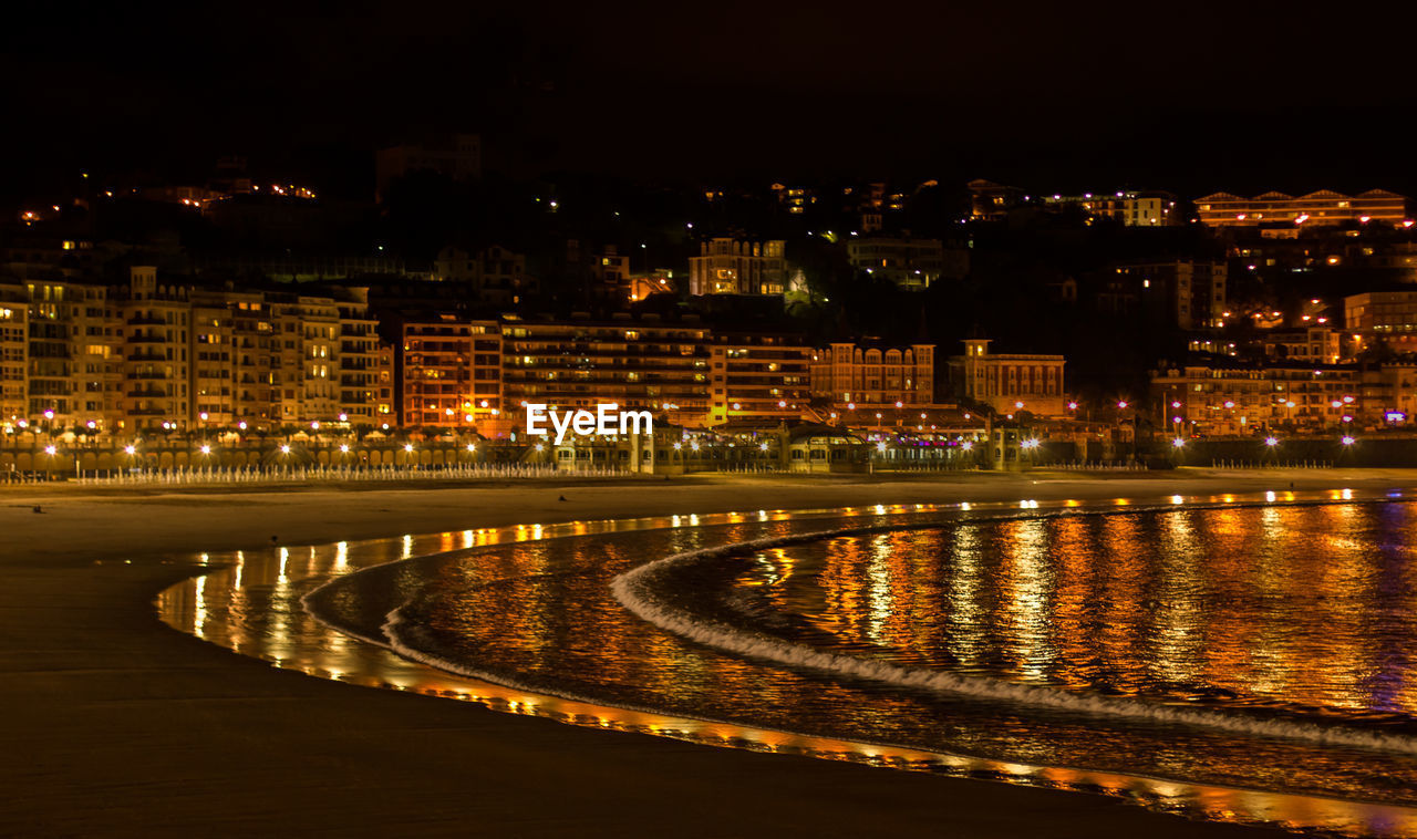 Illuminated buildings by river against sky at night