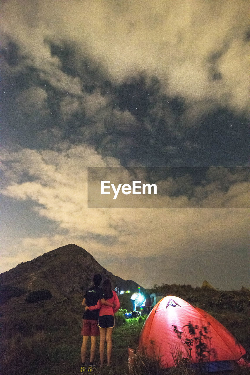 Rear view of man and woman standing by tent on field against cloudy sky at dusk