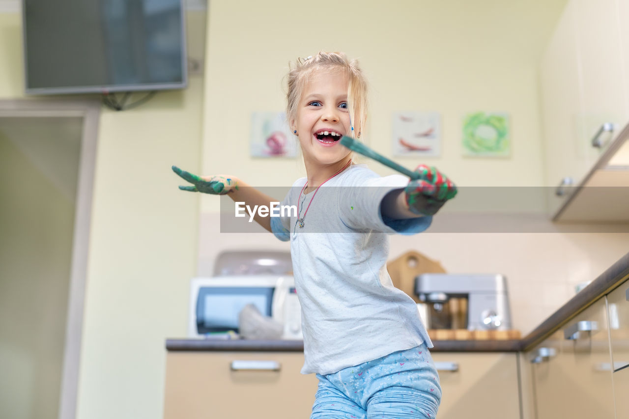 Portrait of cheerful girl holding paintbrush while standing at home