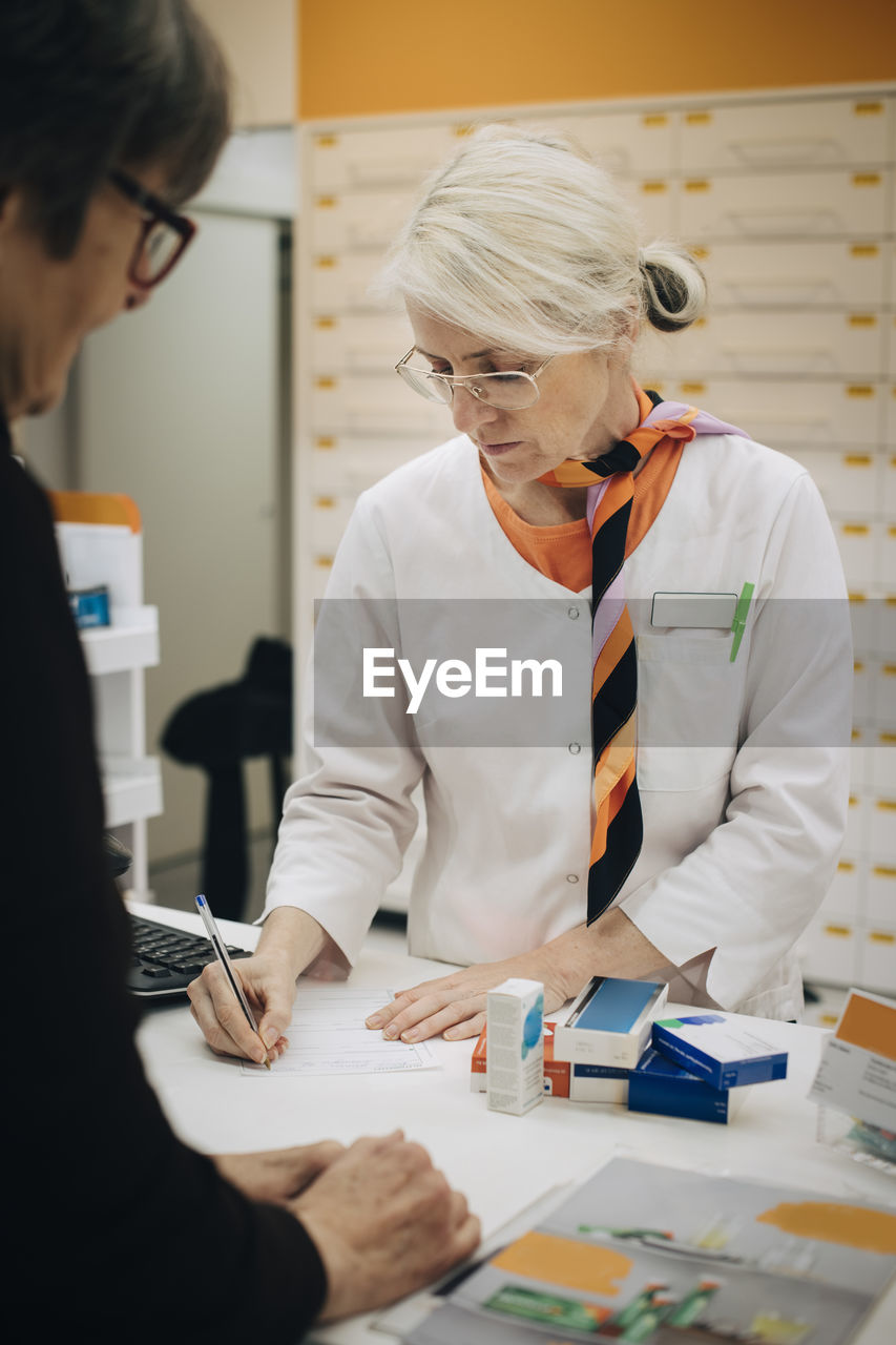 Mature female pharmacist writing on prescription paper at checkout with senior customer in medical store