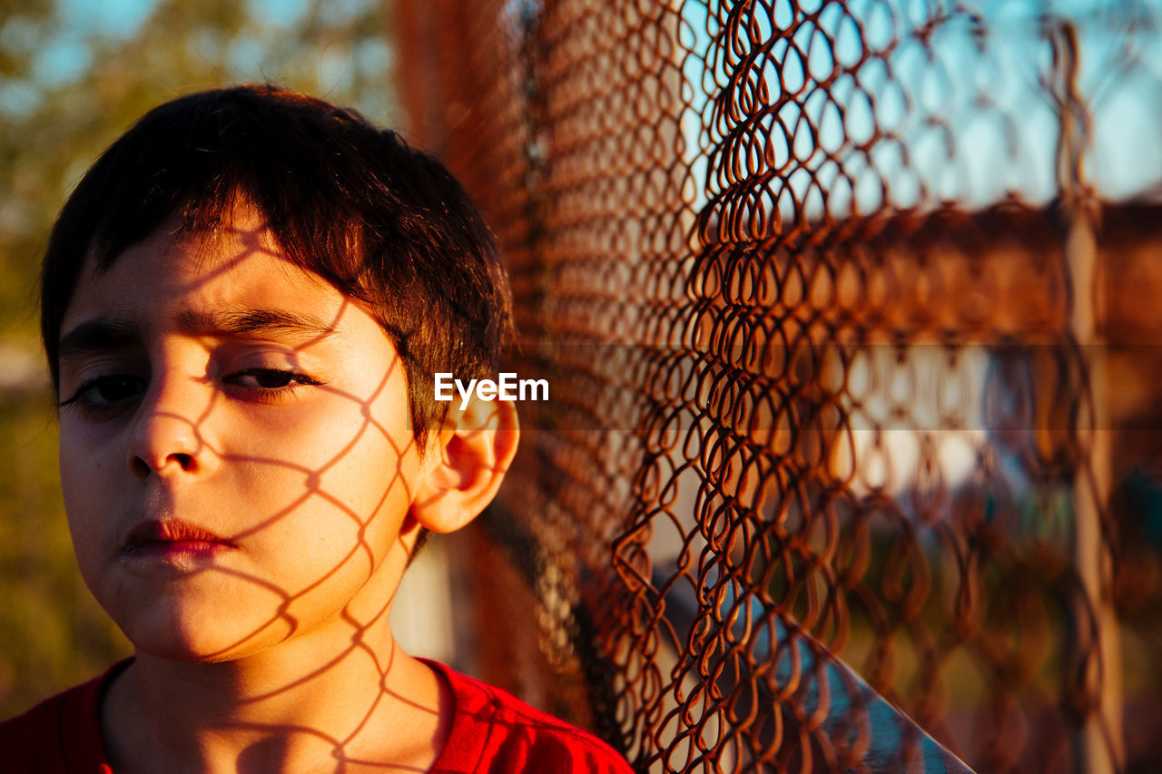 Portrait of boy standing with chain link fence shadow on face outdoors