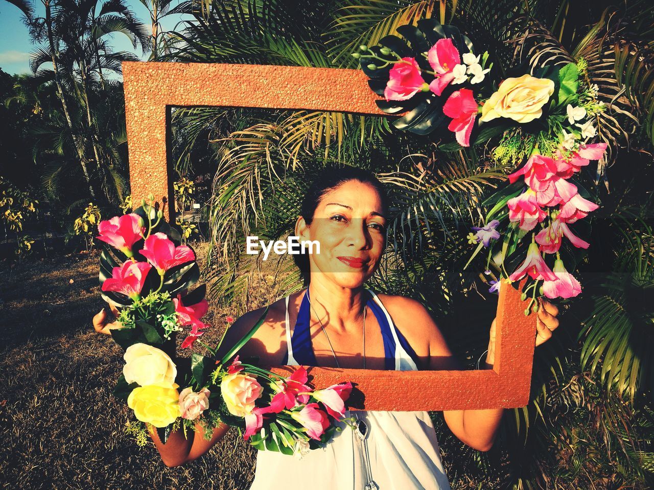 Portrait of mature woman holding picture frame while standing against trees