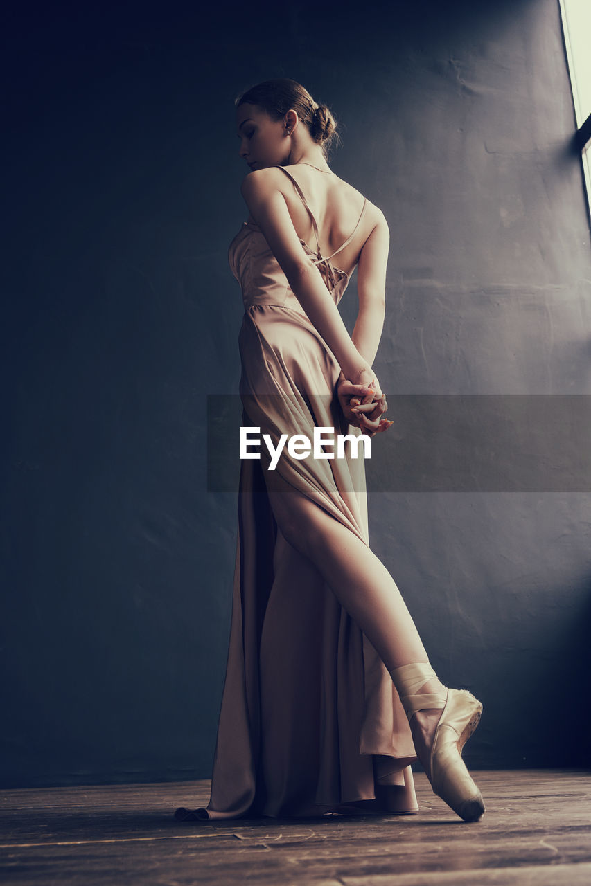 A ballerina in a pink dress and punts is standing at the window with her arms back crossed 