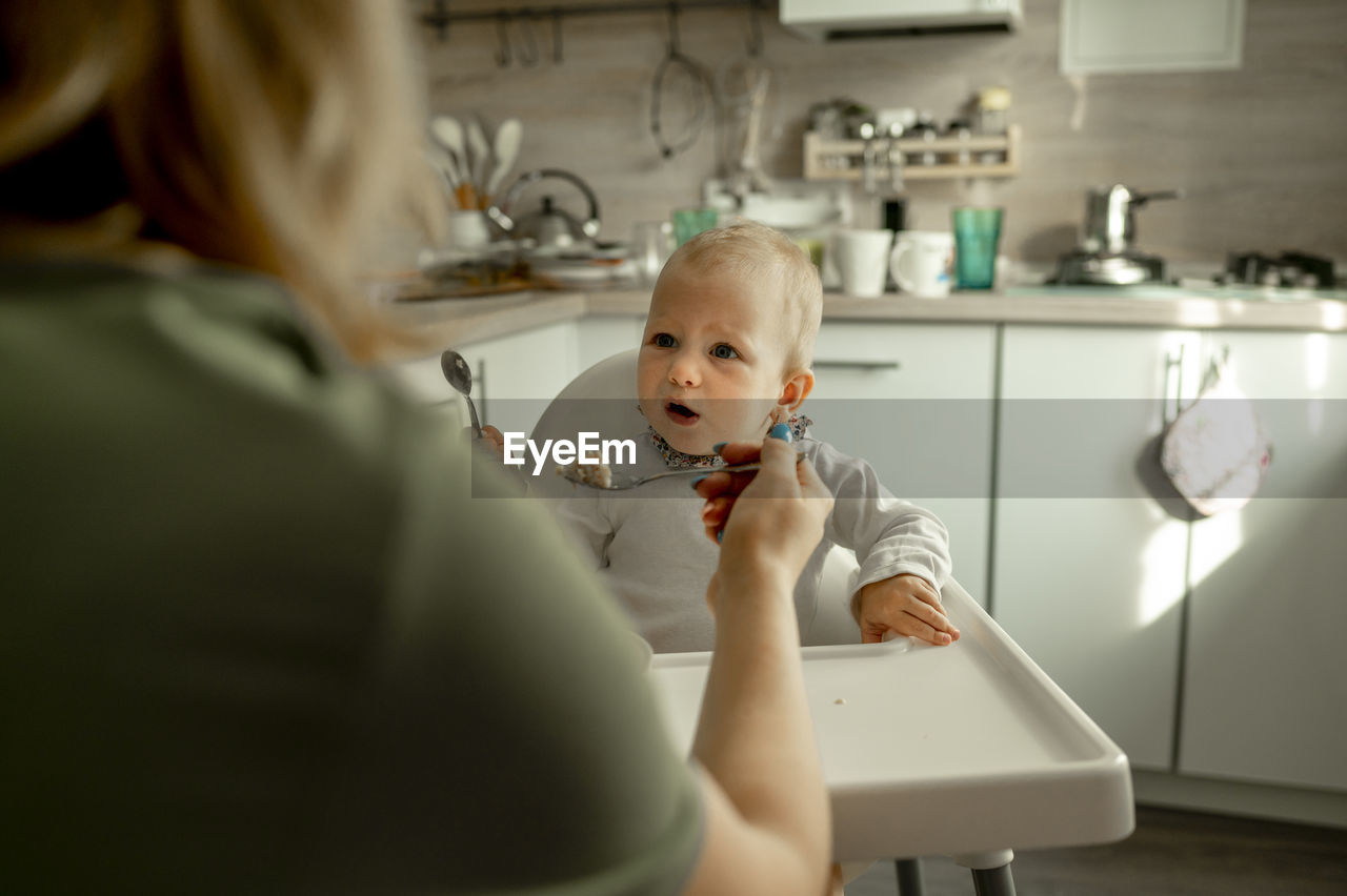 Mother feeding baby girl with porridge in kitchen