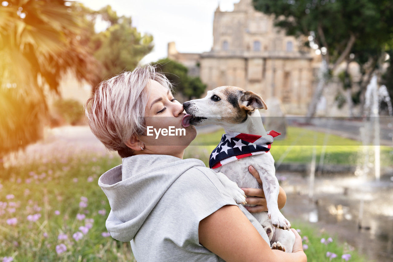 Woman holding small senior dog in park outdoor. dog is wearing bandana with an american flag