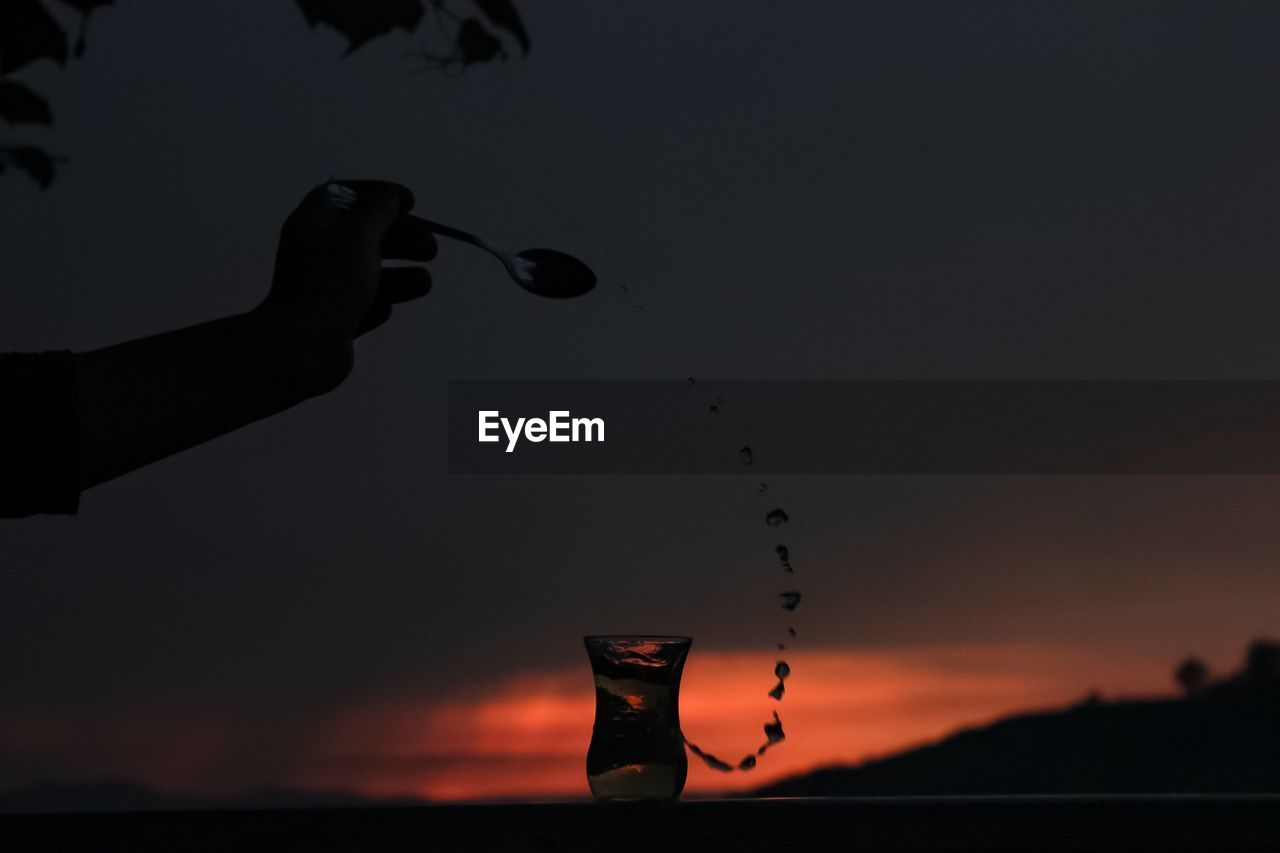 Cropped hand pouring drink in glass against cloudy sky during sunset