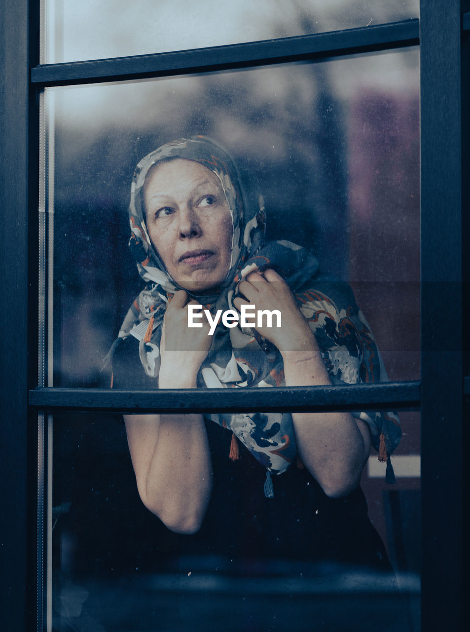 Sad mature woman with scraft on head standing behind dirty window glass