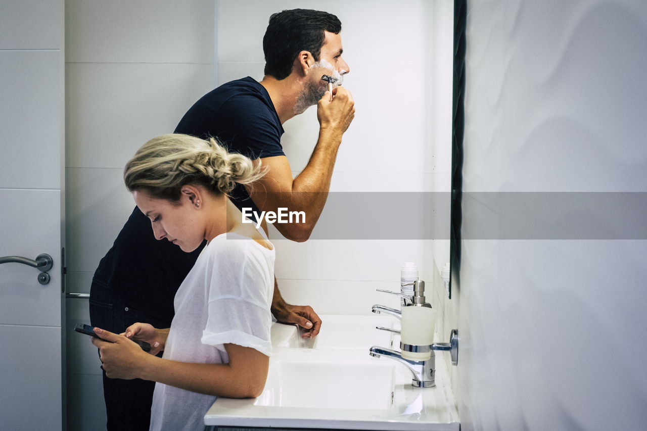 Man shaving with girlfriend using phone in bathroom