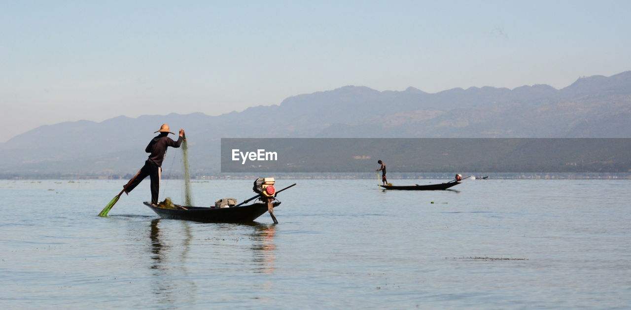 Men fishing while standing on boat in sea against mountains