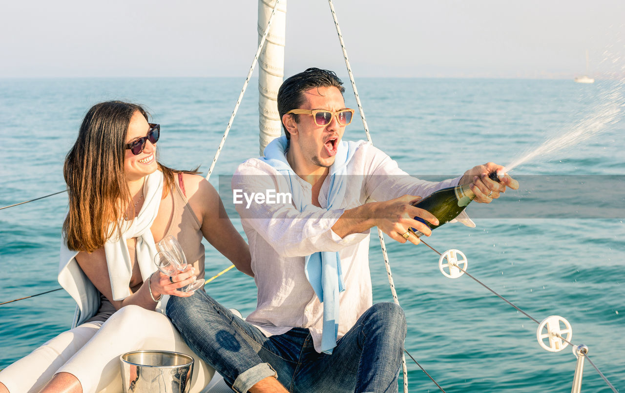 Man spraying champagne with woman holding glasses while sitting on boat in sea