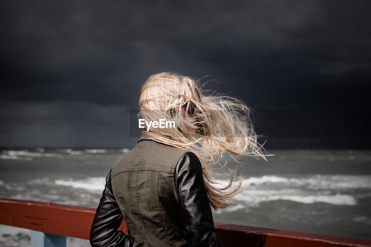 Woman with tousled hair standing by railing at beach against storm clouds