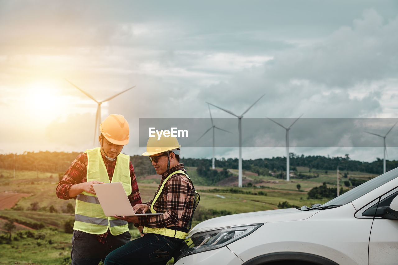 Engineers using laptop by car against cloudy sky