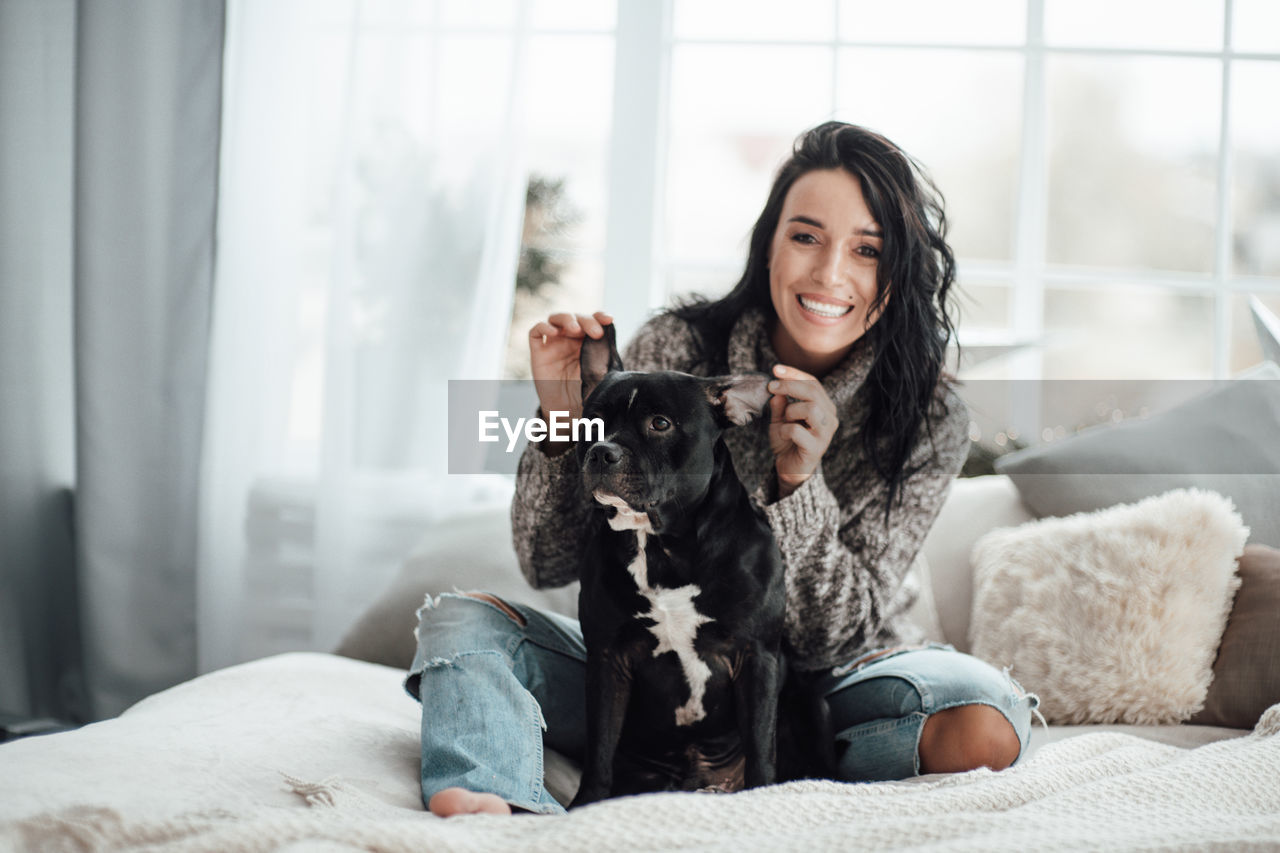 Portrait of smiling woman with dog sitting on bed at home